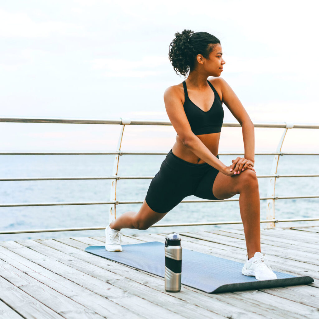 Young woman working out on the pier with a mat and a tumbler of water. 